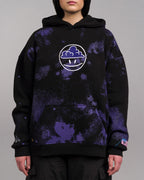 Frontalansicht einer Person mit schwarzem Hoodie. Zentral auf der Brust befindet sich ein weißer, kreisförmiger Print mit einer stilisierten Wolke und zwei Punkten im unteren Bereich. Der Hoodie zeigt ein allover violettes Muster und tropfenförmige Motive am Ärmel sowie einen rosa Label-Patch am Saum.