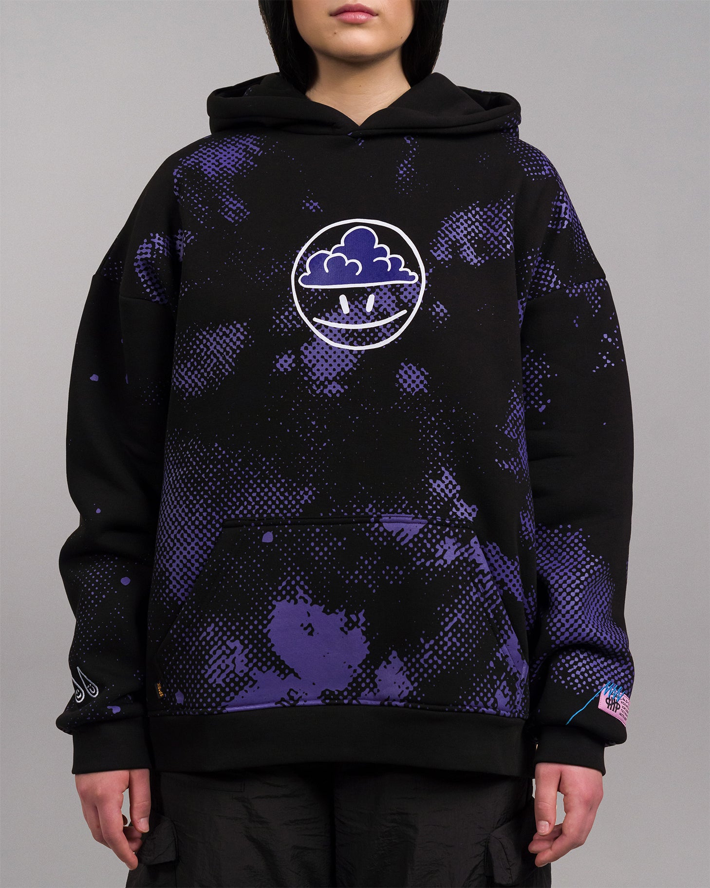 Frontalansicht einer Person mit schwarzem Hoodie. Zentral auf der Brust befindet sich ein weißer, kreisförmiger Print mit einer stilisierten Wolke und zwei Punkten im unteren Bereich. Der Hoodie zeigt ein allover violettes Muster und tropfenförmige Motive am Ärmel sowie einen rosa Label-Patch am Saum.