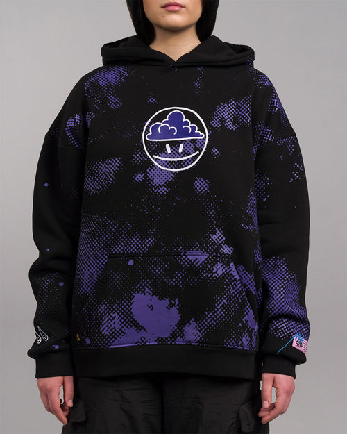 Frontalansicht einer Person mit schwarzem Hoodie. Zentral auf der Brust befindet sich ein weißer, kreisförmiger Print mit einer stilisierten Wolke und zwei Punkten im unteren Bereich. Der Hoodie zeigt ein allover violettes Muster und tropfenförmige Motive am Ärmel sowie einen rosa Label-Patch am Saum.