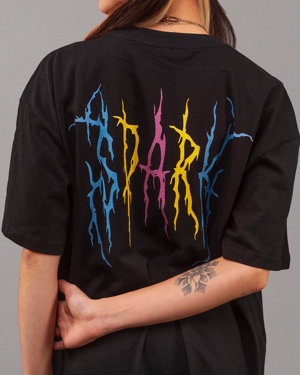 Schwarzes T-Shirt mit großem, farbigem Rückendruck in Flammenoptik. Die Buchstaben des Wortes „SPARK“ sind grafisch verfremdet und erscheinen in den Farben Blau, Gelb und Pink auf dem Rücken des Shirts. Die tätowierte Person trägt das Shirt locker fallend mit zur Seite gedrehter Haltung.