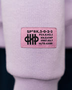Nahaufnahme des lilafarbenen Hoodies zeigt ein rosa Label mit schwarzem Aufdruck auf dem Ärmel. Der Aufdruck enthält typografische Codes und ein grafisches Symbol aus parallelen Linien.