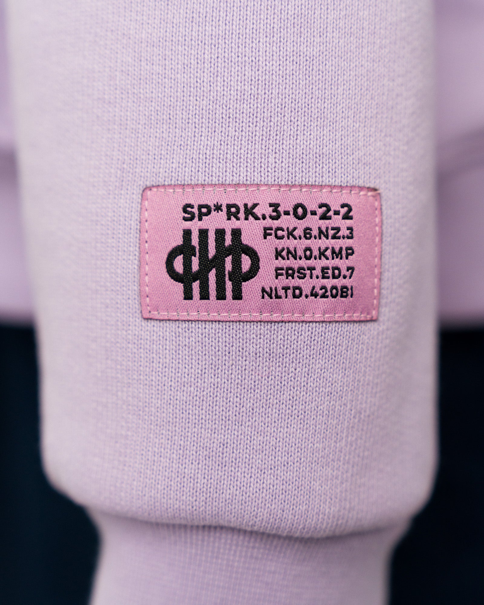 Nahaufnahme des lilafarbenen Hoodies zeigt ein rosa Label mit schwarzem Aufdruck auf dem Ärmel. Der Aufdruck enthält typografische Codes und ein grafisches Symbol aus parallelen Linien.