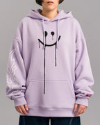Vorderansicht des lilafarbenen Hoodies an einer stehenden Person. Die Brust ist mit einem großen schwarzen Smiley bedruckt, dessen Augen und Mund verlaufen. Linker Ärmel mit weißem Flammenprint und rosa Label über der rechten Bündchennaht.