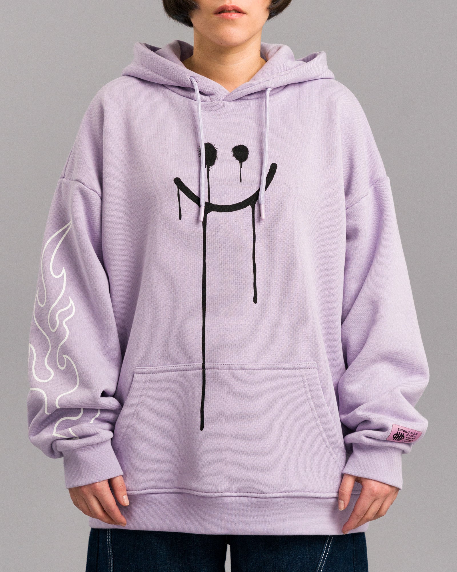 Vorderansicht des lilafarbenen Hoodies an einer stehenden Person. Die Brust ist mit einem großen schwarzen Smiley bedruckt, dessen Augen und Mund verlaufen. Linker Ärmel mit weißem Flammenprint und rosa Label über der rechten Bündchennaht.