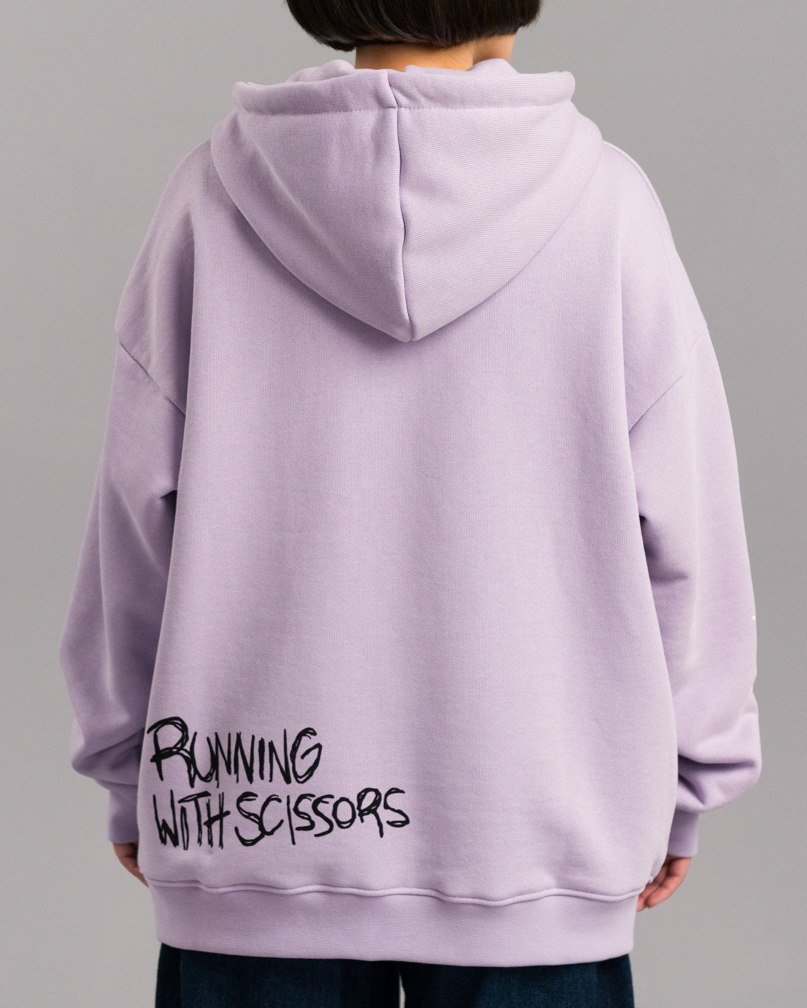 Rückansicht des lilafarbenen Hoodies mit schwarzem, handschriftlich wirkendem Schriftzug „RUNNING WITH SCISSORS“ im unteren Rückenbereich.