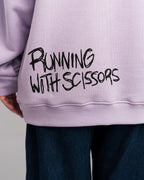 Detailaufnahme des Rückendrucks mit der Aufschrift „RUNNING WITH SCISSORS“ in schwarzer, handgemalter Schrift auf fliederfarbenem Hoodie.