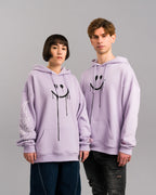Zwei Personen tragen den lilafarbenen Hoodie mit schwarzem Smileyprint. Die linke Person zeigt den Ärmel mit weißem Flammenmotiv, beide Hoodies haben rosa Label auf dem Ärmel.