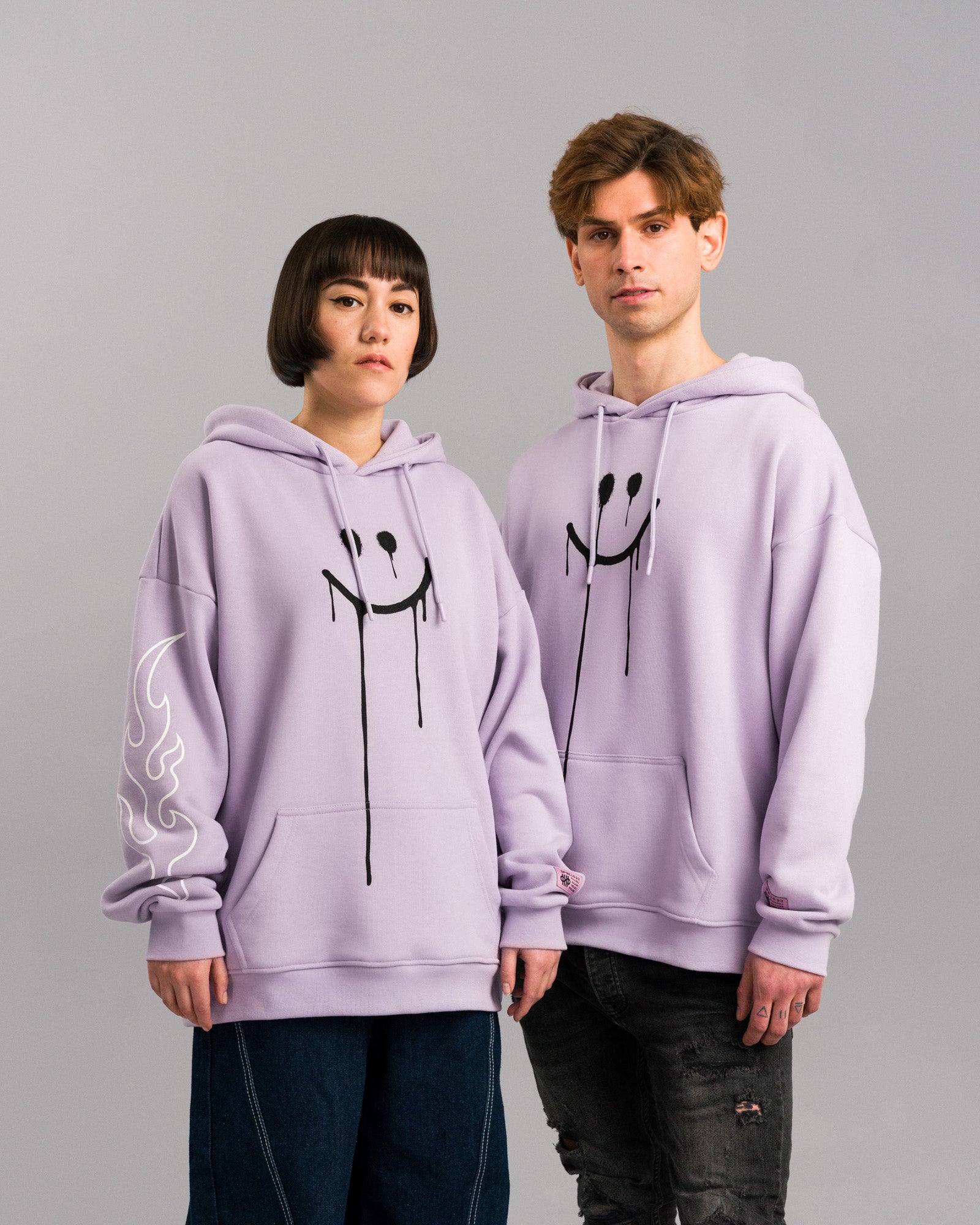 Zwei Personen tragen den lilafarbenen Hoodie mit schwarzem Smileyprint. Die linke Person zeigt den Ärmel mit weißem Flammenmotiv, beide Hoodies haben rosa Label auf dem Ärmel.