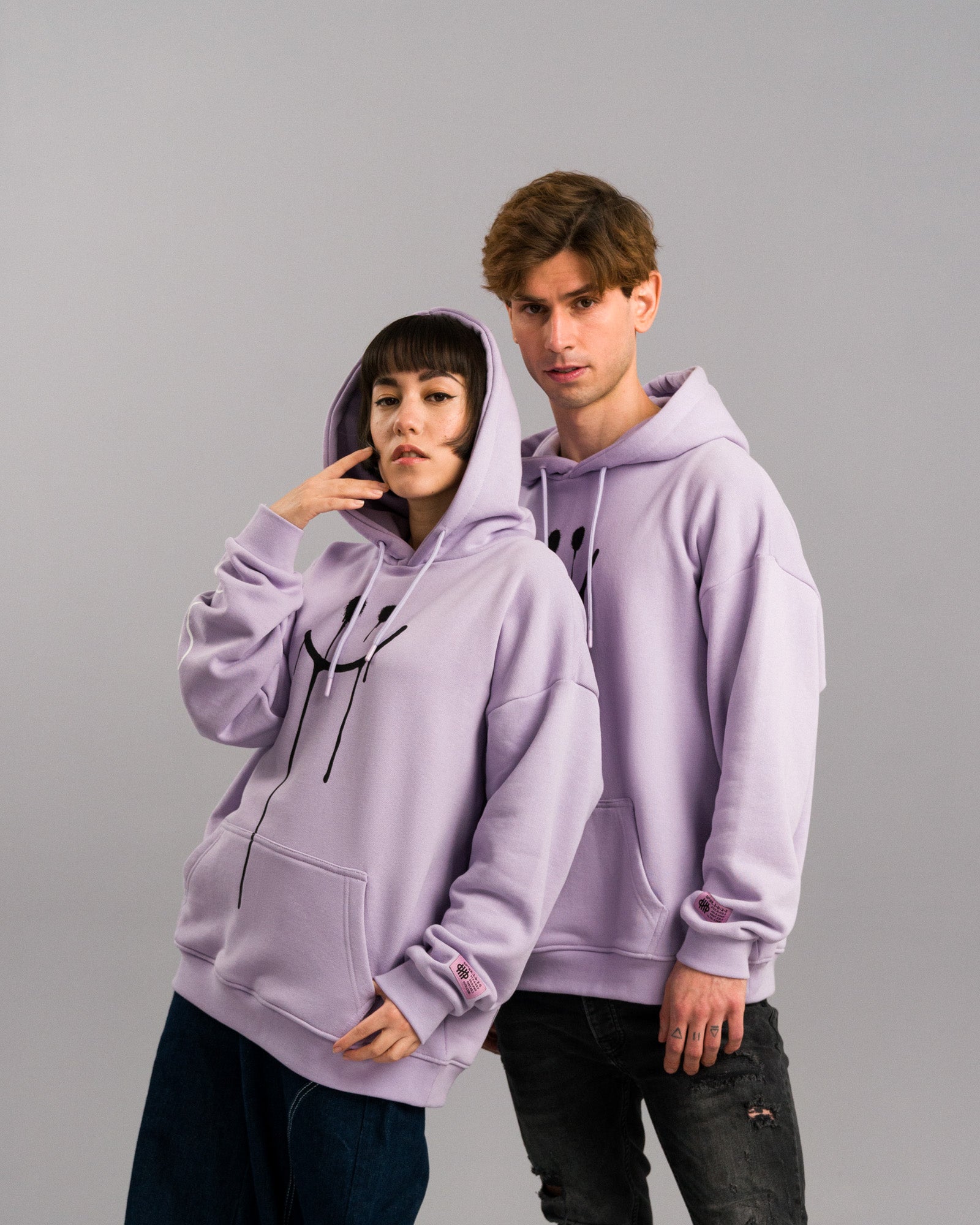 Zwei Personen in identischen lilafarbenen Hoodies mit aufgesetzter Kapuze. Der Smileyprint und das rosa Ärmel-Label sind sichtbar, eine Person hebt die Hand ans Gesicht.