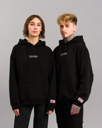 Zwei Personen tragen den gleichen schwarzen Hoodie mit pastellfarbener SP/RK-Stickerei und pinkem Label auf dem linken Ärmel.