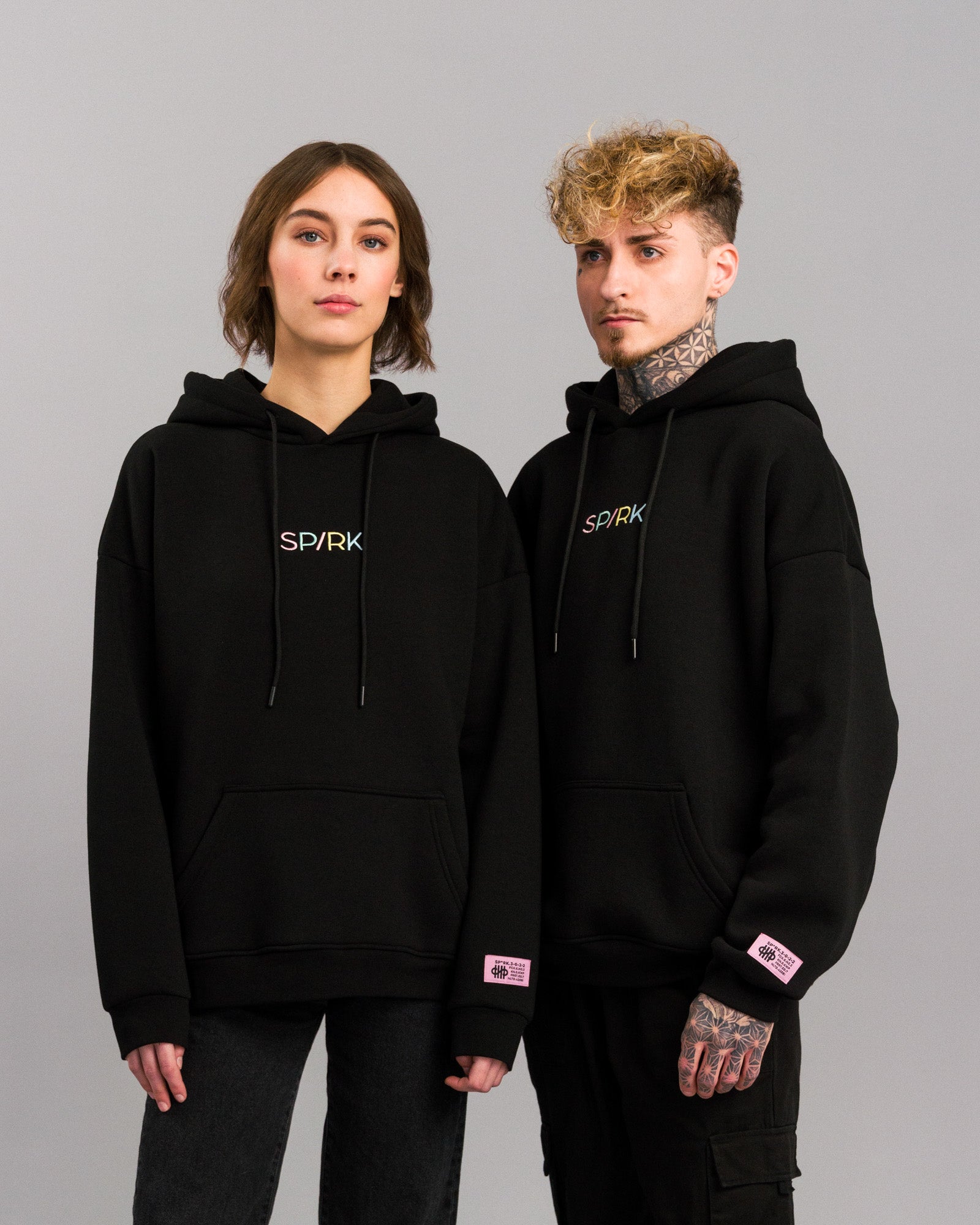 Zwei Personen tragen den gleichen schwarzen Hoodie mit pastellfarbener SP/RK-Stickerei und pinkem Label auf dem linken Ärmel.