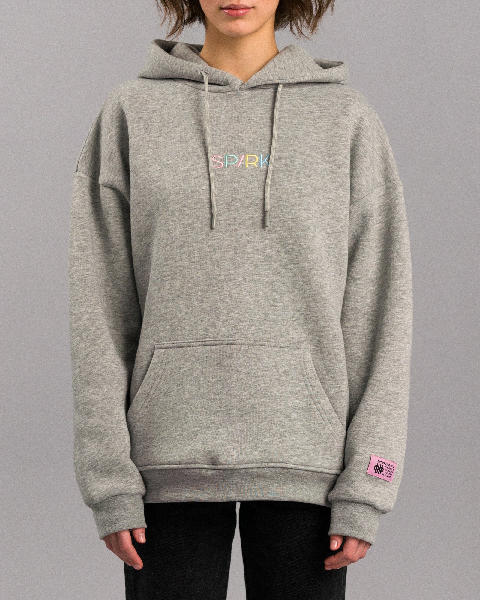 Grauer Kapuzenpullover in lockerer Passform mit Kängurutasche und verstellbarer Kapuze. Auf der Brust ist der Schriftzug „SP/RK“ in bunten Pastellfarben gestickt. Am Ärmelabschluss befindet sich ein kleines rosa Label mit schwarzem Text und grafischen Elementen.