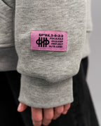 Detailansicht des rosa Labels am Ärmel des grauen Hoodies. Das Label zeigt schwarze Zahlen- und Buchstabencodes sowie ein grafisches Logo mittig.
