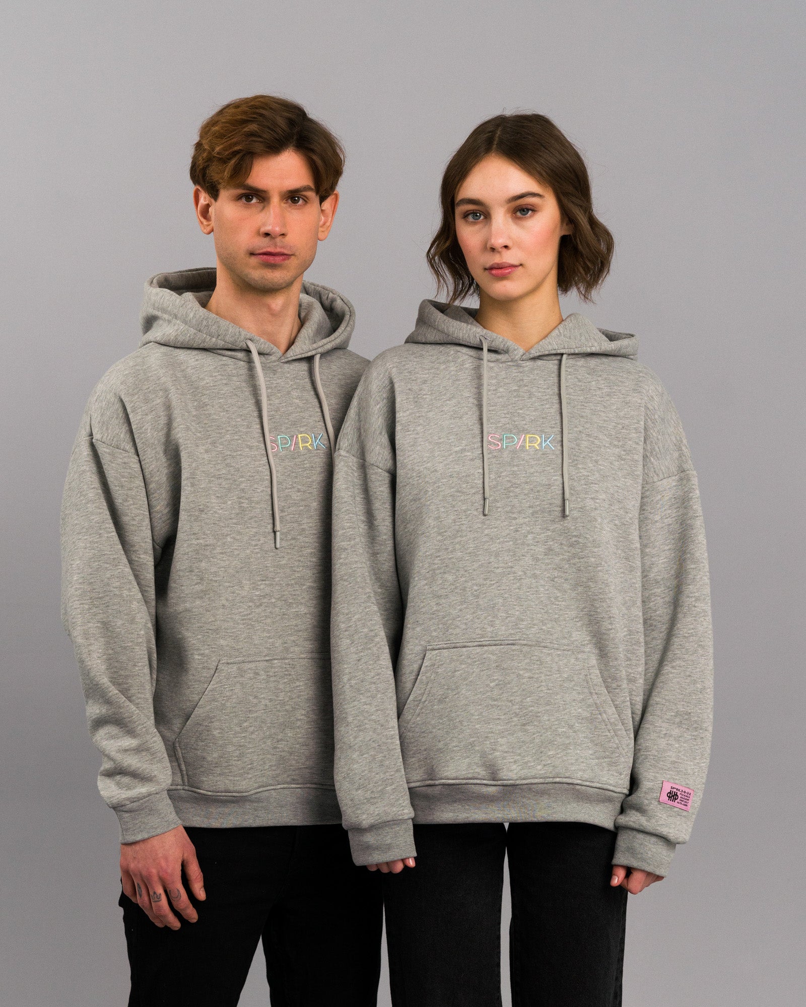 Zwei Personen tragen denselben grauen Kapuzenpullover mit buntem „SP/RK“-Bruststick. Beide Modelle stehen nebeneinander vor neutralem Hintergrund, das linke Modell mit sichtbarem Label am rechten Ärmel.