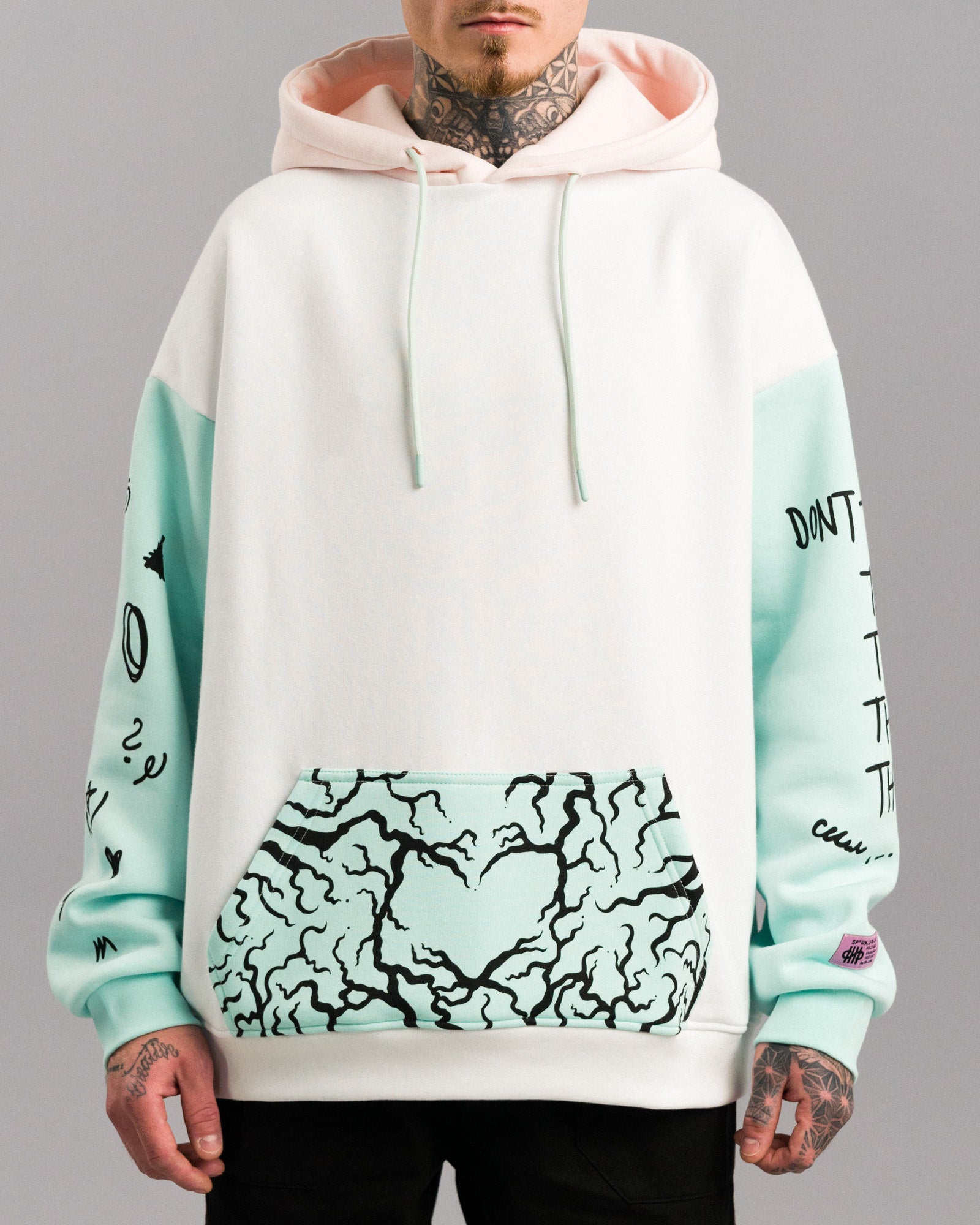 Hoodie in Weiß mit mintfarbenen Ärmeln und Kängurutasche. Die Tasche ist mit einem schwarzen, verästelten Herzmuster bedruckt. Die Kapuze ist innen rosa gefüttert. Auf dem linken Ärmel sind schwarze Illustrationen verteilt, auf dem rechten Ärmel beginnt eine Typografie.
