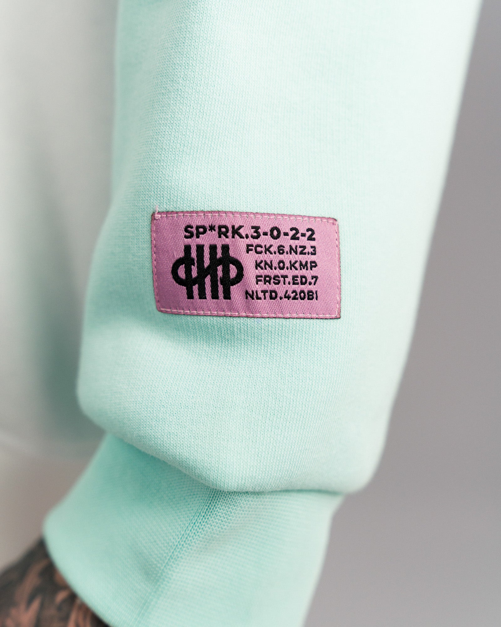 Nahaufnahme des lilafarbenen Labelpatches auf dem rechten Ärmel mit kryptischen Textzeilen und dem stilisierten Logo.
