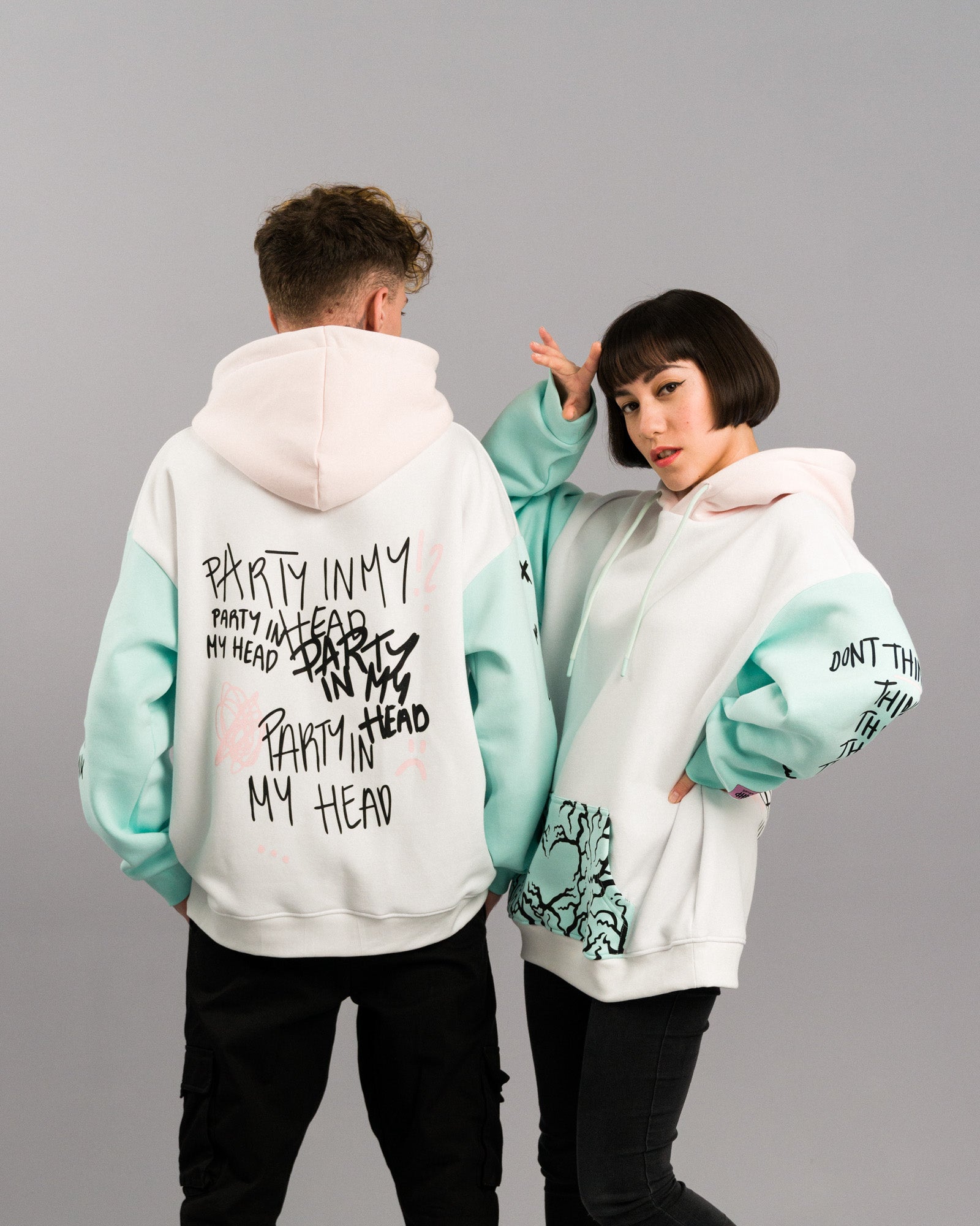 Beide Models in dynamischer Pose. Die Rückseite des linken Hoodies zeigt den „PARTY IN MY HEAD“-Print, der rechte Hoodie ist leicht seitlich gedreht und zeigt Vorderseite mit Tasche und bedrucktem Ärmel.