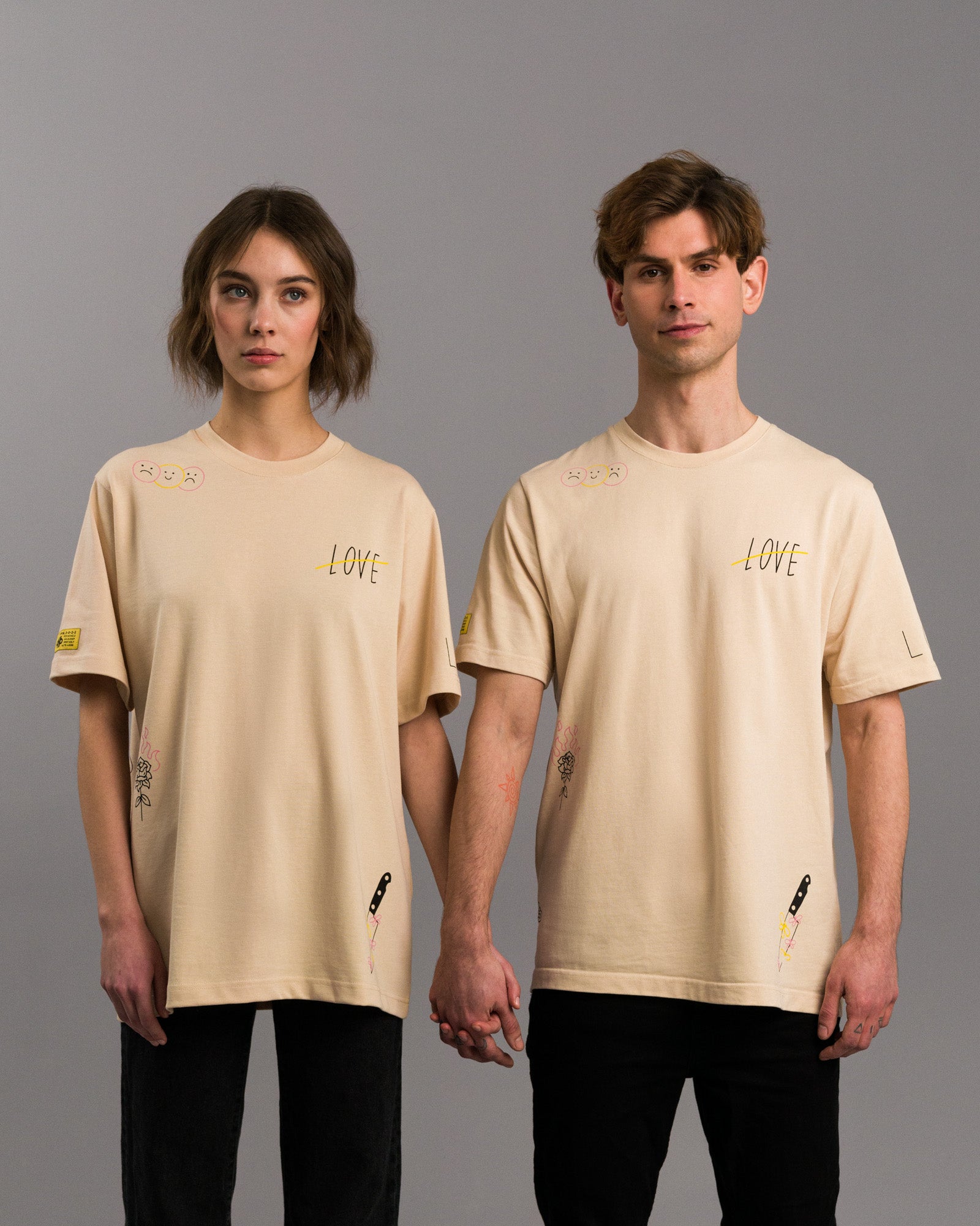 Zwei Personen in identischem cremefarbenem T-Shirt mit mehreren grafischen Illustrationen, darunter ein durchgestrichenes „LOVE“, ein Messer mit Blumen, eine brennende Rose und kleine Smileys. Beide stehen frontal nebeneinander und halten sich an den Händen.