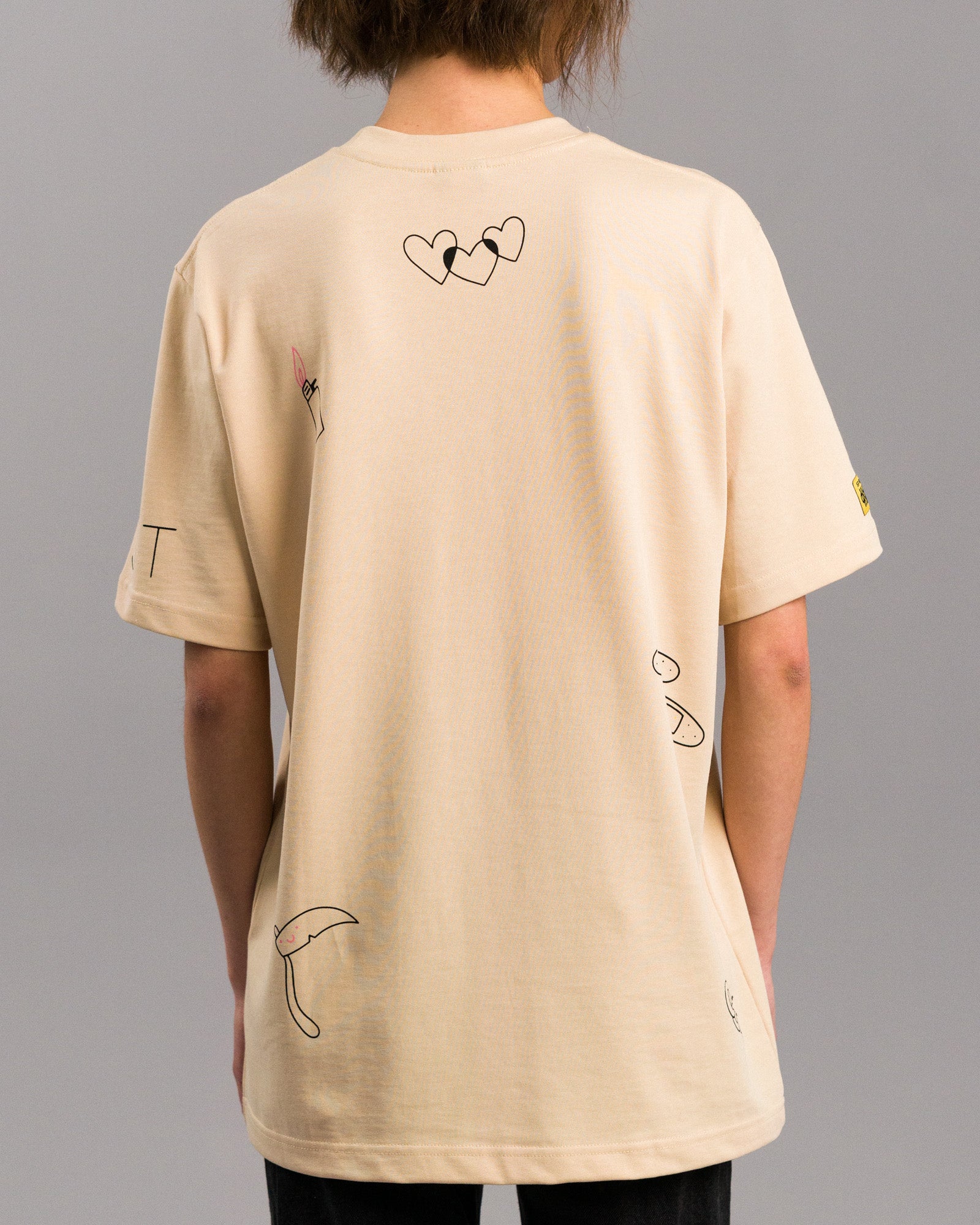 Rückansicht des T-Shirts mit mehreren grafischen Motiven: drei sich überlappende Herzen im oberen Bereich, ein kleiner Lippenstift links, eine Sense unten sowie weitere abstrakte Symbole auf der rechten Seite.