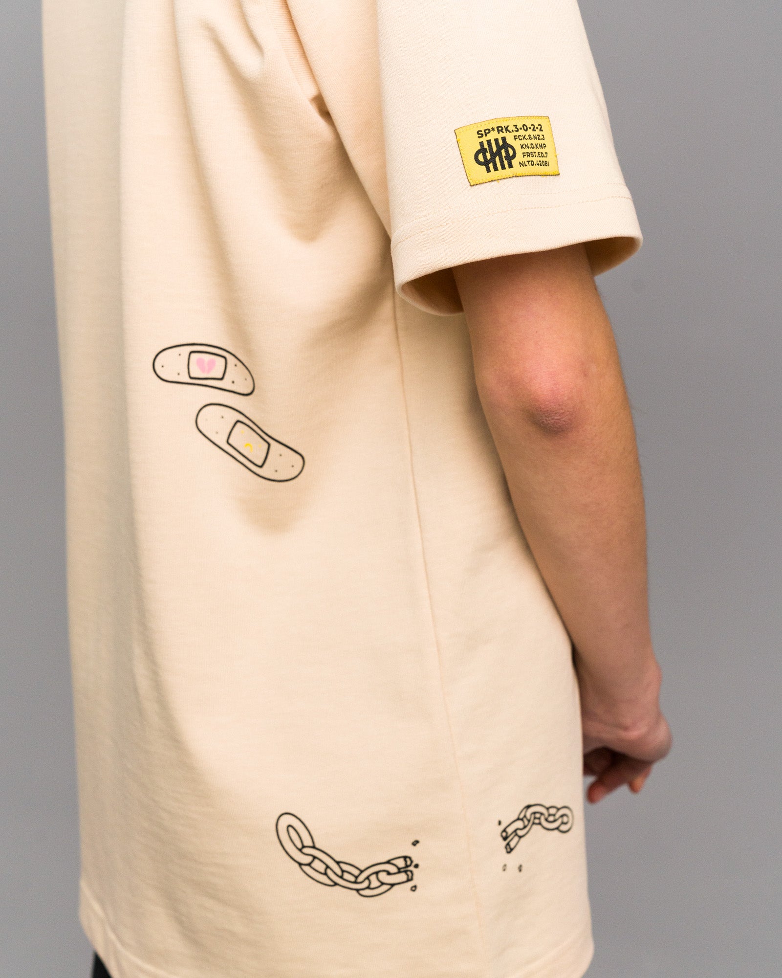 Seitliche Detailaufnahme mit Fokus auf eine zwei Pflaster mit Herz-Icons und eine zerbrochene Kette auf der unteren Rückseite. Das gelbe Label ist auf dem linken Ärmel zu sehen.