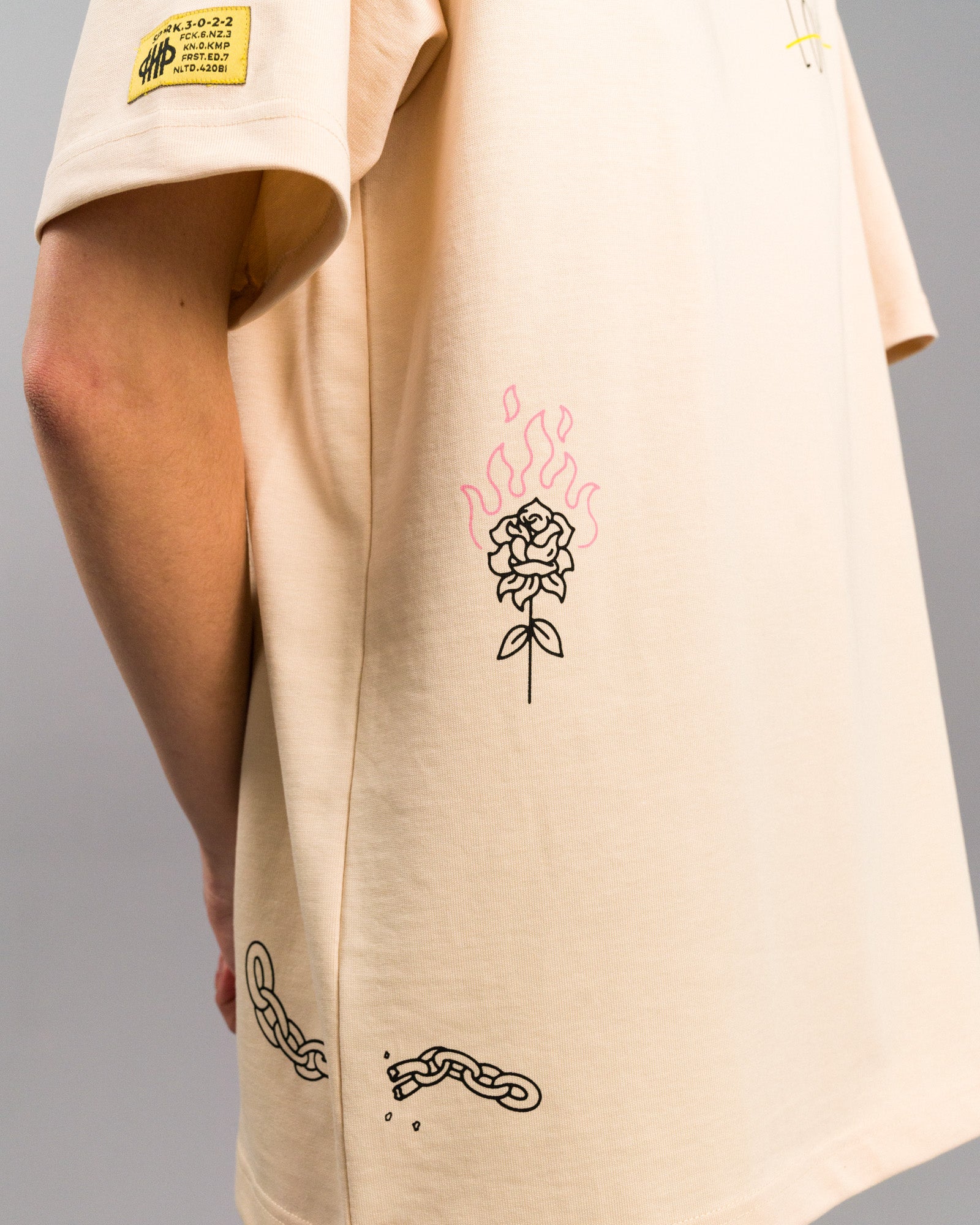Seitliche Detailaufnahme mit Fokus auf eine brennende Rose und eine zerbrochene Kette auf der unteren Rückseite. Das gelbe Label ist auf dem linken Ärmel zu sehen.