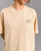 Nahaufnahme eines cremefarbenen T-Shirts mit schwarzem „LOVE“-Schriftzug auf der Brust, der durch eine gelbe Linie durchgestrichen ist. Die linke Schulter zeigt drei Smiley-Gesichter, zwei traurig, eines fröhlich. Getragen von einer Person vor grauem Hintergrund.