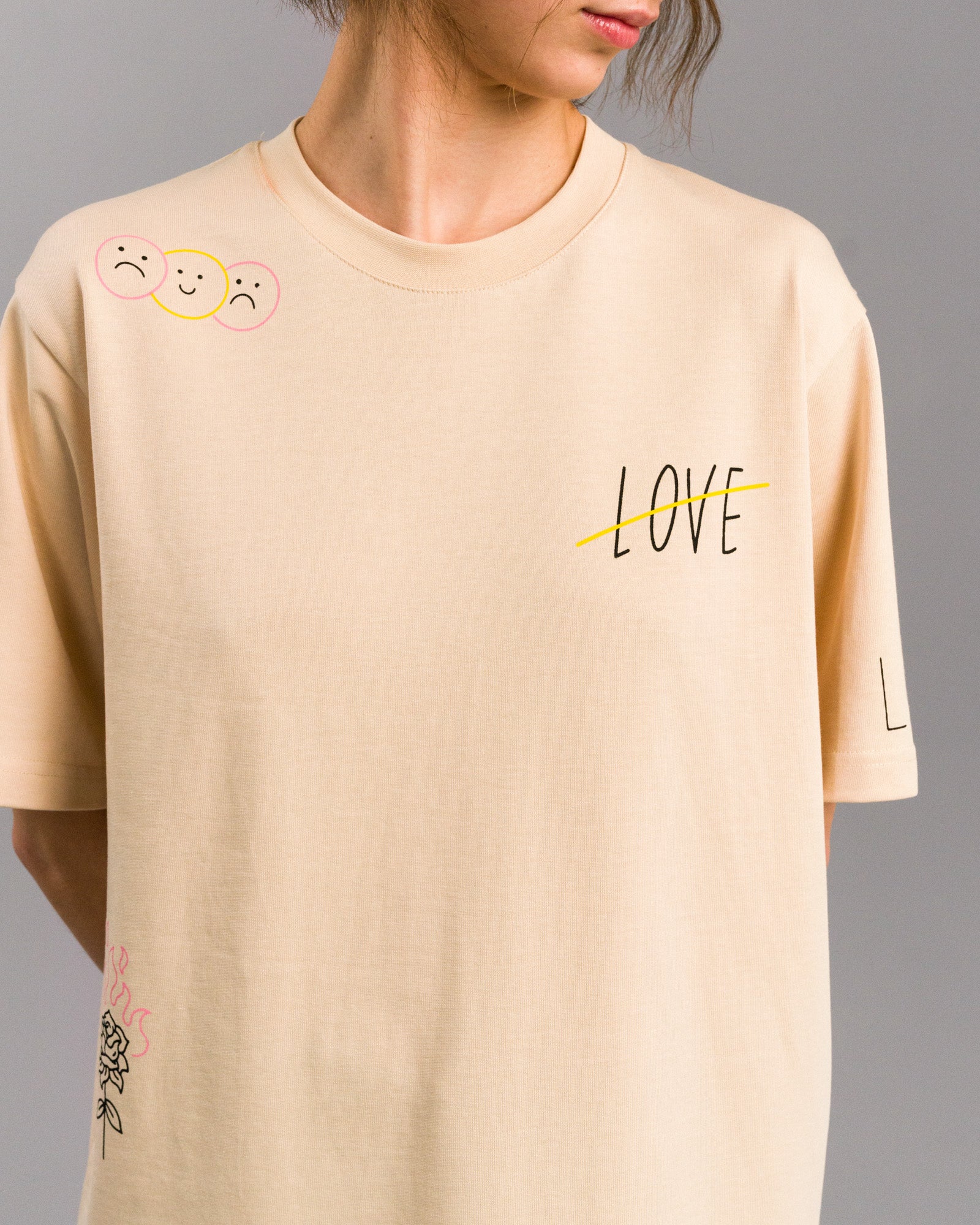 Nahaufnahme eines cremefarbenen T-Shirts mit schwarzem „LOVE“-Schriftzug auf der Brust, der durch eine gelbe Linie durchgestrichen ist. Die linke Schulter zeigt drei Smiley-Gesichter, zwei traurig, eines fröhlich. Getragen von einer Person vor grauem Hintergrund.