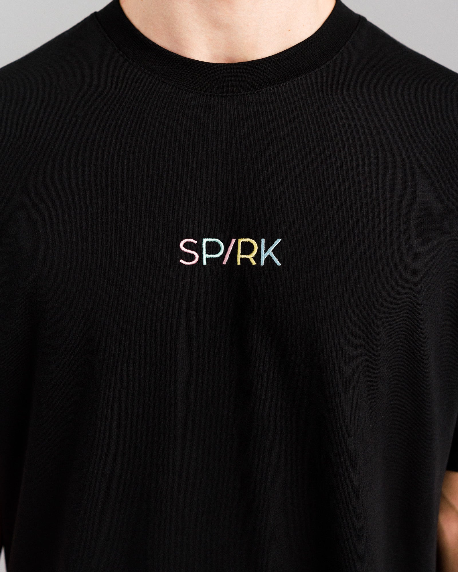 Detailansicht des bunten, mittig platzierten Schriftzugs „SPARK“ auf der Brust des schwarzen Shirts.