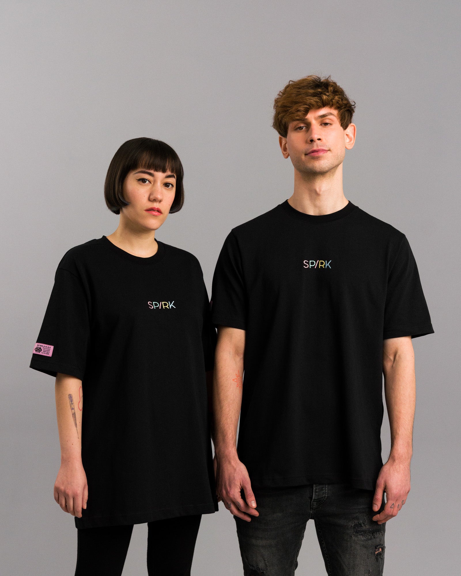 Zwei Personen tragen das schwarze SPARK-T-Shirt mit farbigem Logo und Label am linken Ärmel, locker geschnitten.