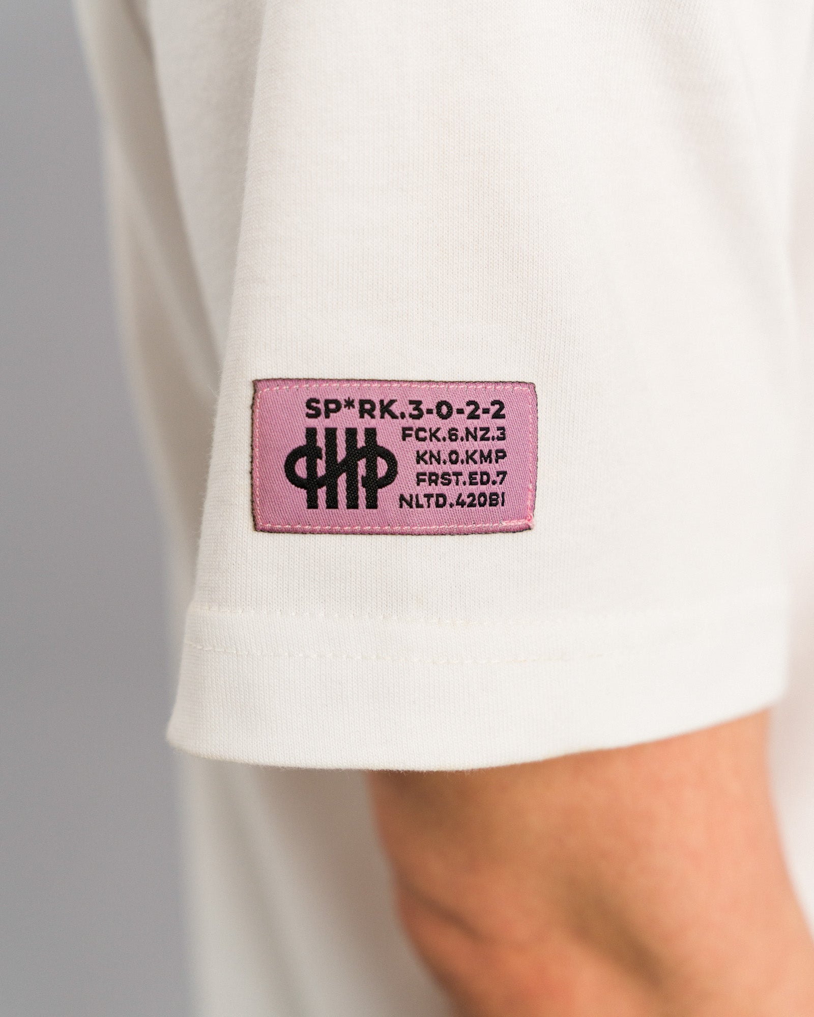Linker Ärmel des weißen T-Shirts mit aufgenähtem pinkfarbenem Label, bedruckt mit schwarzen Symbolen und codiertem Text.