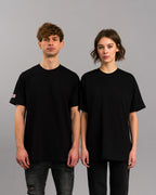 Zwei Personen tragen das schwarze SP/RK-T-Shirt mit Ton-in-Ton-Stickerei und pinkem Labelpatch, beide in Frontalansicht stehend.