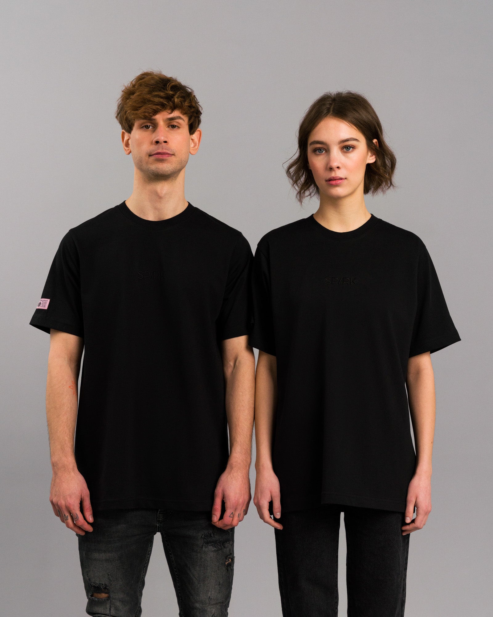 Zwei Personen tragen das schwarze SP/RK-T-Shirt mit Ton-in-Ton-Stickerei und pinkem Labelpatch, beide in Frontalansicht stehend.