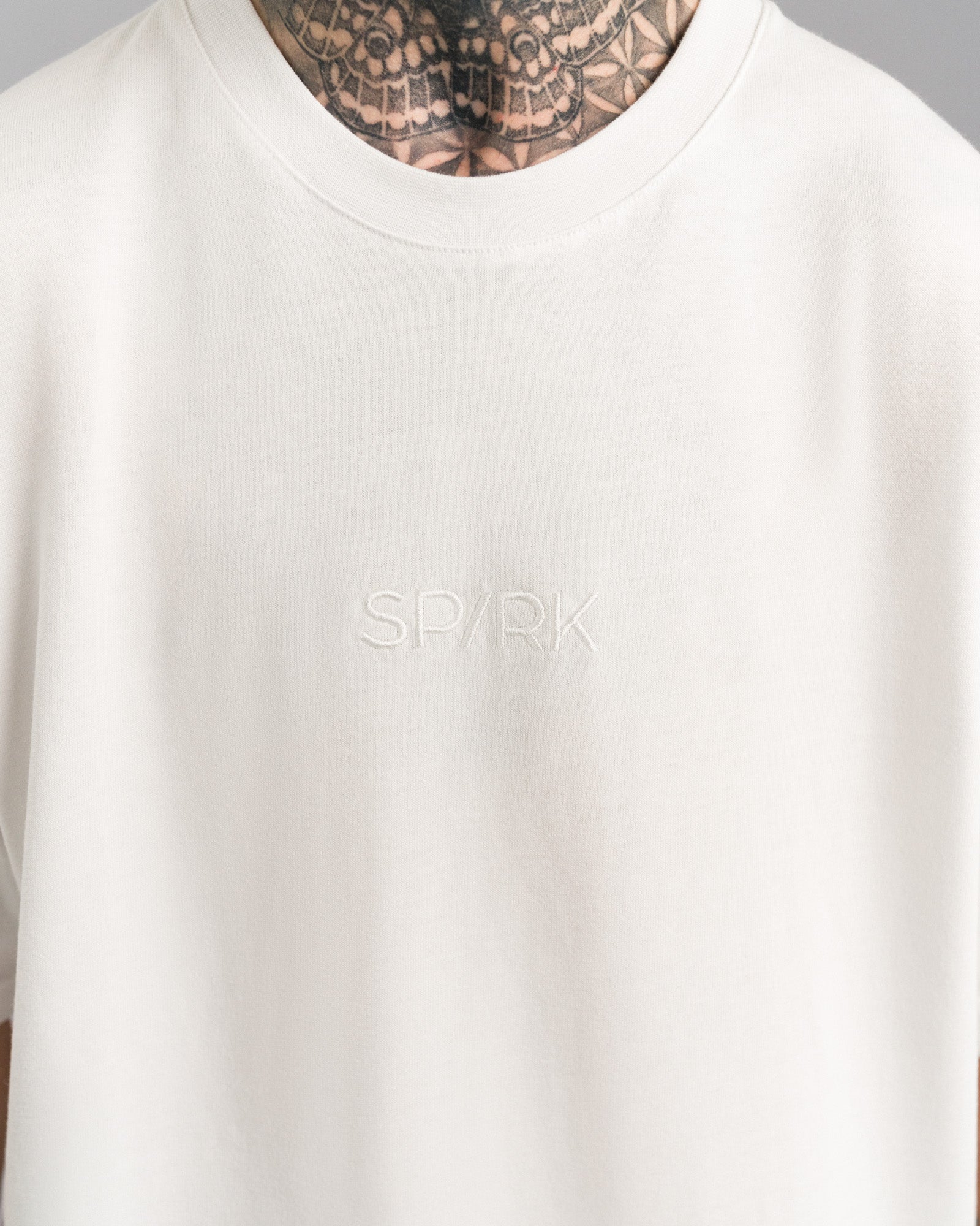 Nahaufnahme der weißen „SPARK“-Stickerei auf der Brust des weißen T-Shirts, Ton-in-Ton gestaltet.