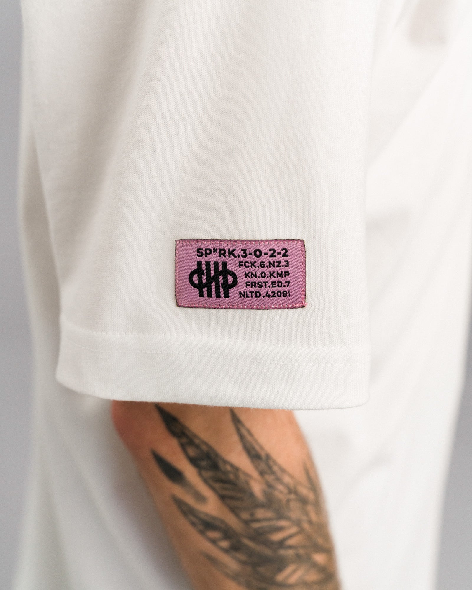 Detailaufnahme des linken Ärmels mit pinkfarbenem, eckigem Label mit schwarzem Text und Logo auf dem weißen Stoff.