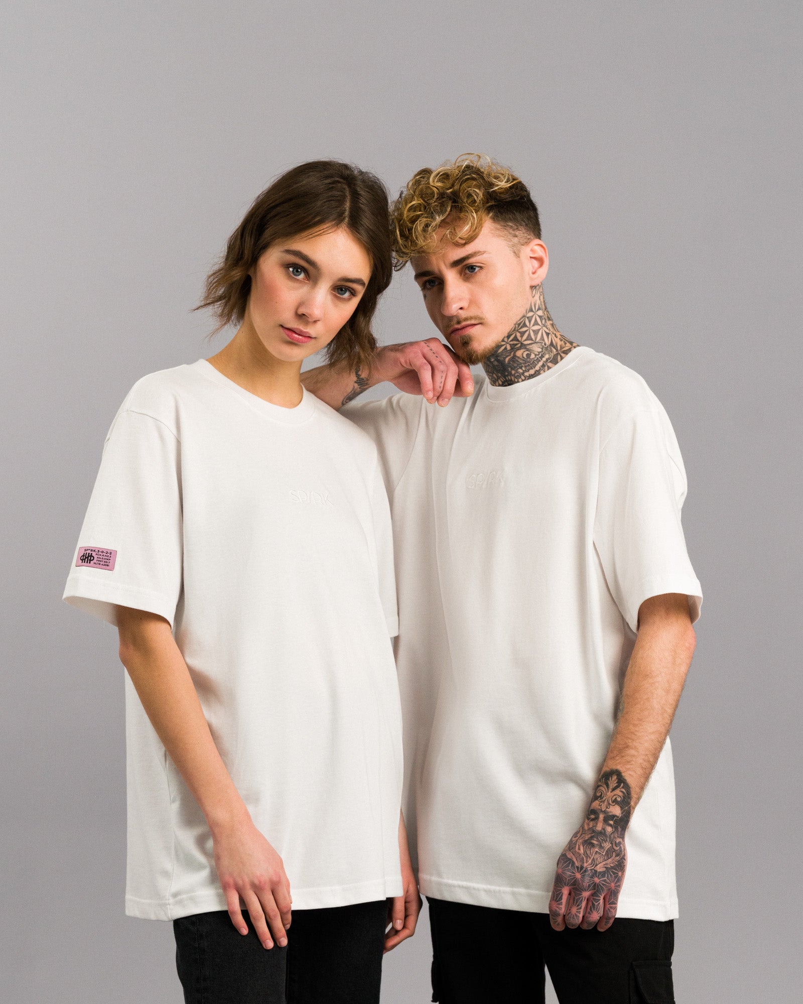 Zwei Personen (eine weiblich, ein männlich) tragen das weiße T-Shirt mit weißem „SPARK“-Stick auf der Brust. Beide stehen eng beieinander vor grauem Hintergrund.