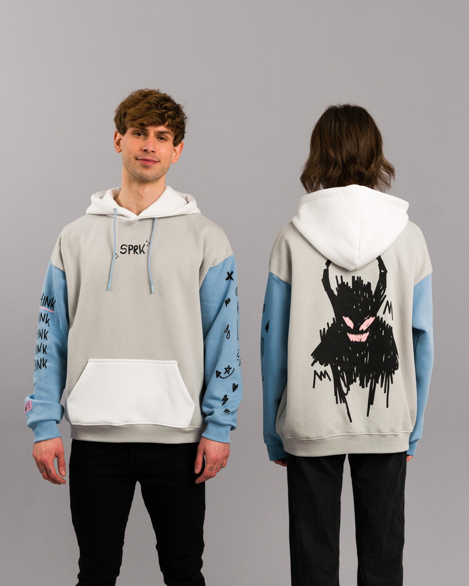 Zwei Personen tragen denselben Hoodie, jeweils von vorne und von hinten gezeigt. Der Hoodie ist hellgrau mit hellblauen Ärmeln und weißer Kapuze. Auf der Rückseite befindet sich eine große schwarze Kritzelgrafik eines dämonenartigen Wesens mit pinken Augen und Mund. Der linke Ärmel zeigt wiederholt den Schriftzug „THINK“ sowie weitere Illustrationen.