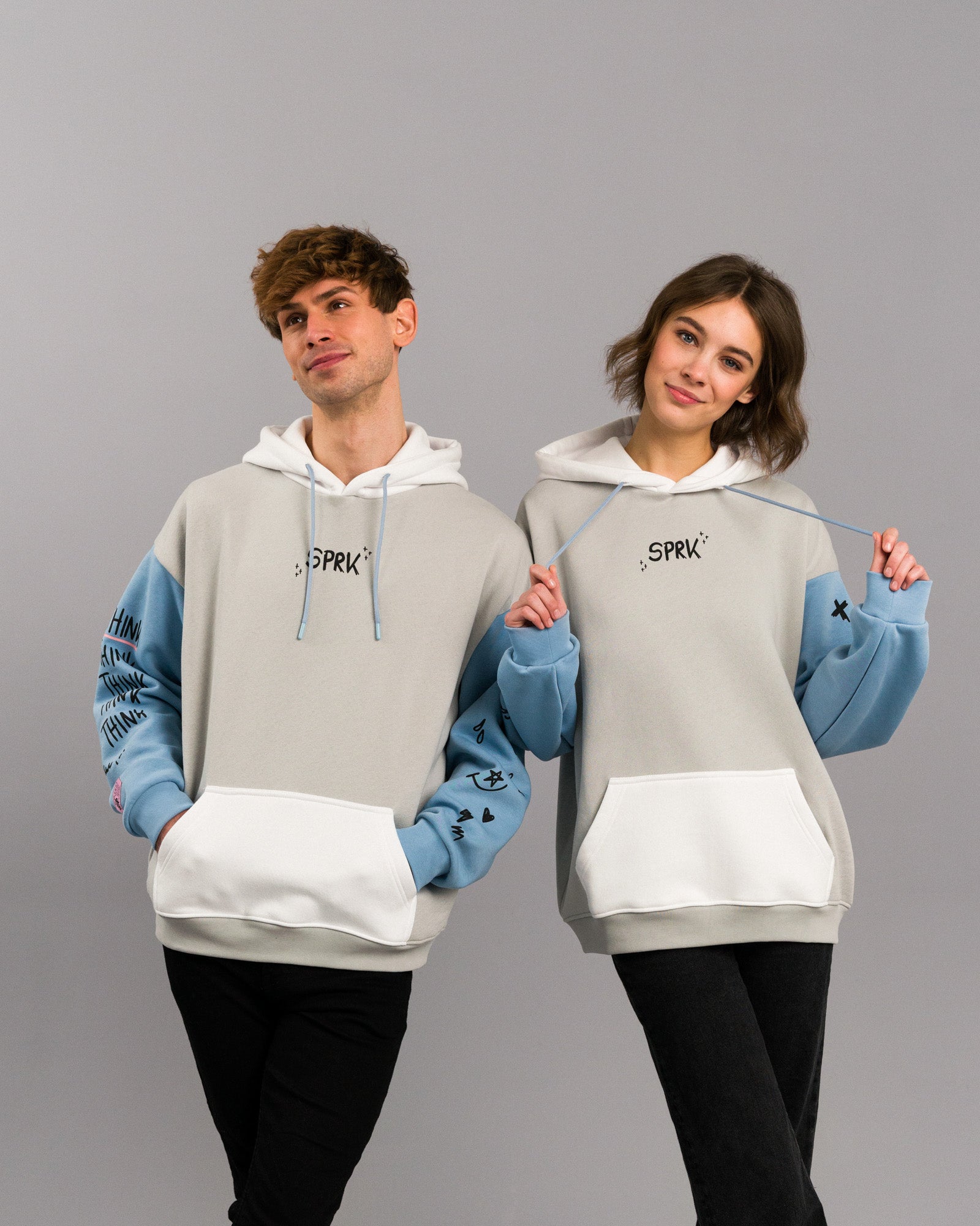 Zwei Personen stehen nebeneinander und tragen identische Hoodies mit hellem Grundton, weißen Kapuzen und hellblauen Ärmeln. Auf der Brust befindet sich ein kleiner „SPRK“-Schriftzug, auf den Ärmeln verschiedene Symbole und der Schriftzug „THINK“. Beide Personen lächeln und posieren spielerisch.