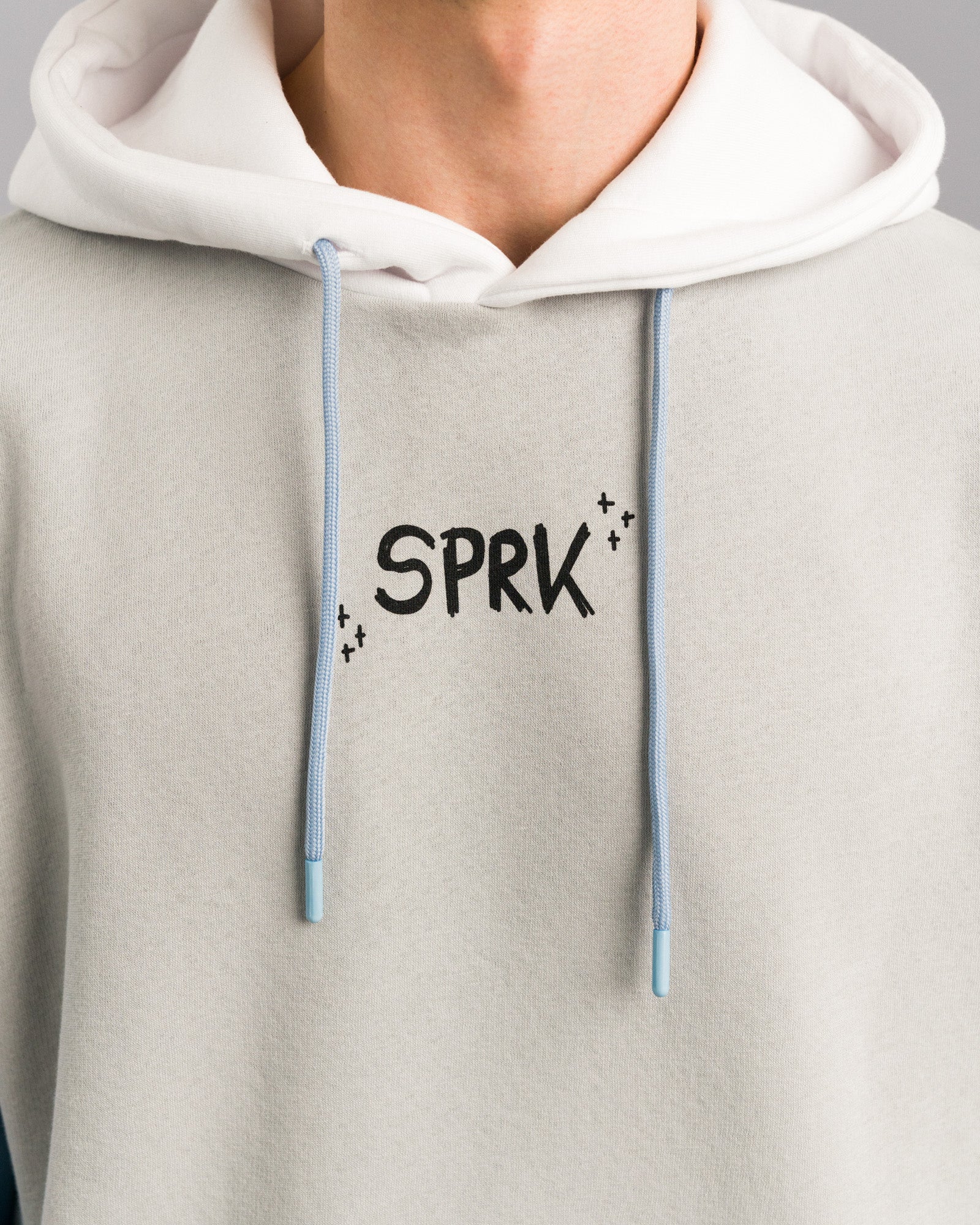 Nahaufnahme des oberen Hoodie-Bereichs. Zu sehen ist der schwarze Schriftzug „SPRK“ auf der Brust sowie der weiße Kapuzenbereich mit hellblauen Kordeln.