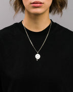 Schwarzes T-Shirt mit silberfarbener Halskette, getragen von einer Person. Die Halskette besteht aus einem flachen Gliederkettenband und einem runden Anhänger mit einem grafischen Smiley-Gesicht in Schwarz, dessen Mund und Augen in verlaufender Tropfenform gestaltet sind.

