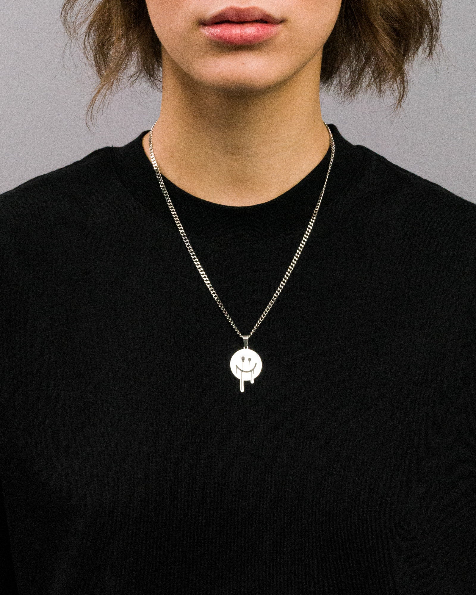 Schwarzes T-Shirt mit silberfarbener Halskette, getragen von einer Person. Die Halskette besteht aus einem flachen Gliederkettenband und einem runden Anhänger mit einem grafischen Smiley-Gesicht in Schwarz, dessen Mund und Augen in verlaufender Tropfenform gestaltet sind.

