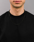 Nahaufnahme einer schwarzen Panzerkette aus Edelstahl, getragen über einem schwarzen T-Shirt. Die gleichmäßig geschwungenen Glieder liegen flach am Hals an und erzeugen ein glänzendes, markantes Finish.