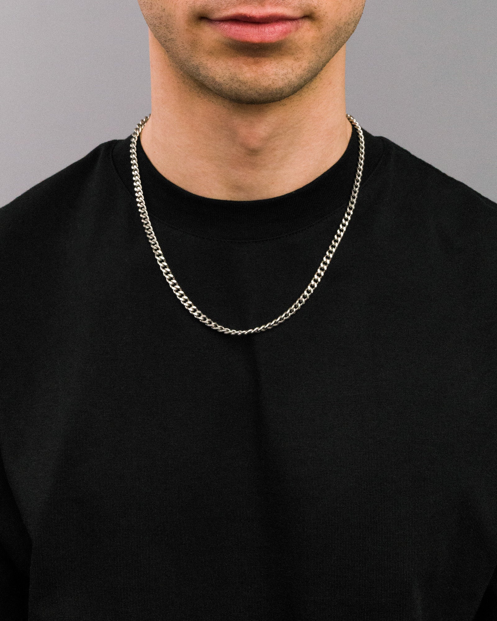 Nahaufnahme einer silberfarbenen Gliederkette in mittelbreitem Design, getragen über einem schwarzen T-Shirt. Die Kette liegt locker um den Hals und hebt sich kontrastreich vom dunklen Hintergrund ab.