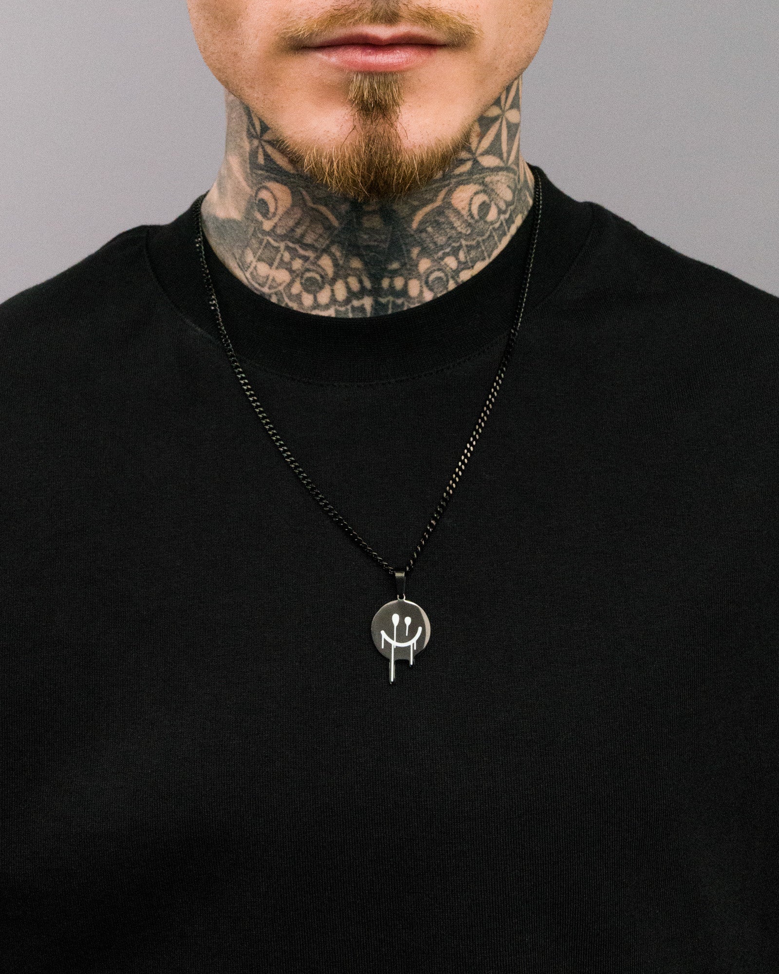 Nahaufnahme einer schwarzen Halskette mit rundem, schwarzem Anhänger. Der Anhänger zeigt einen weißen Smiley mit verlaufenden Linien, getragen zu einem schwarzen T-Shirt.