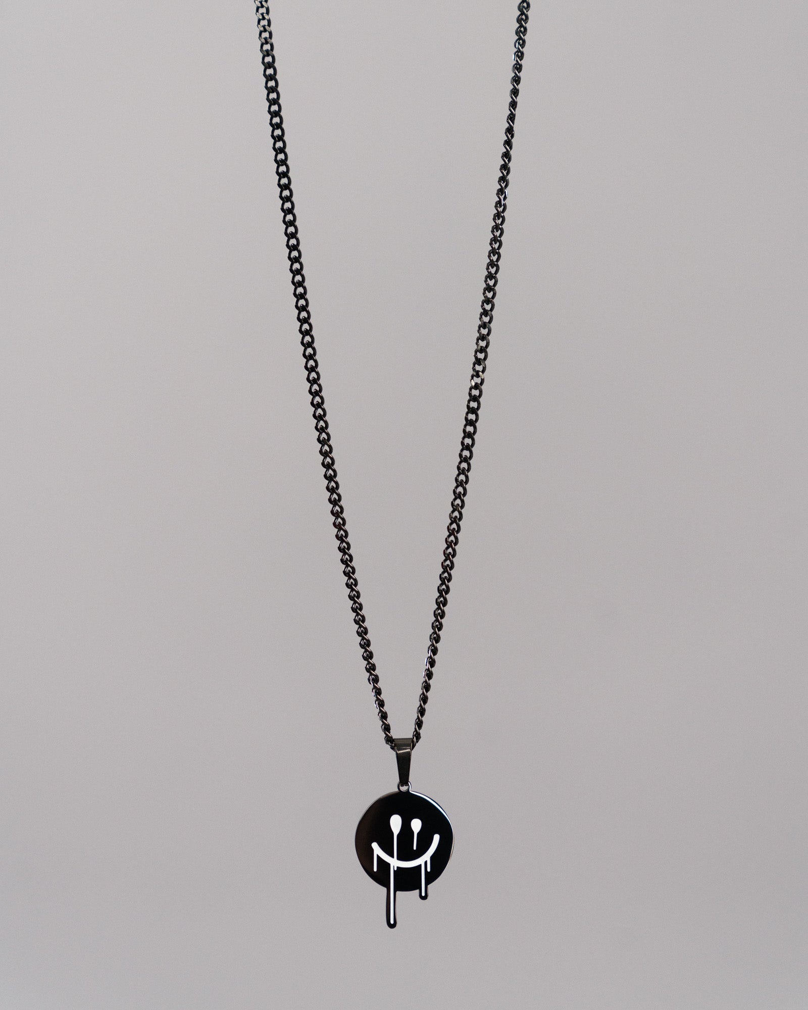 Freigestellte Aufnahme einer schwarzen Halskette mit rundem Anhänger vor grauem Hintergrund. Der Anhänger zeigt ein weißes Smiley-Gesicht mit herunterlaufenden Linien, passend zum Hoodie-Design.

