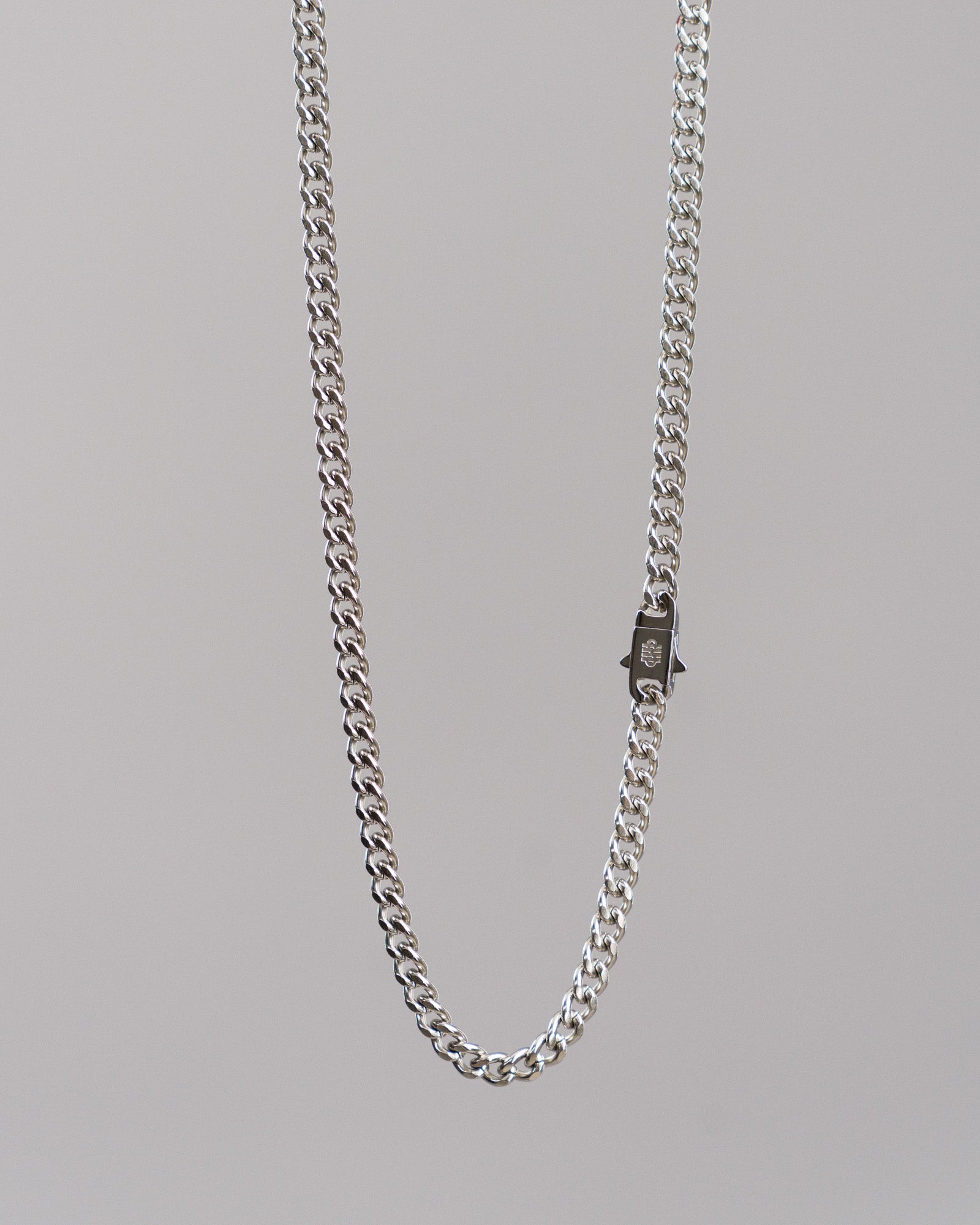 Freigestellte silberfarbene Gliederkette mit mittlerer Gliederstärke vor hellem Hintergrund. Die Kette zeigt ein kleines, rechteckiges Verschlussdetail mit Logo-Gravur in der Mitte der Aufnahme.