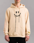 Beigefarbener Hoodie mit schwarzem Spray-Print eines tropfenden Smileys auf der Vorderseite, getragen von einer Person. Der Hoodie besitzt eine Kängurutasche und eine Kapuze mit Kordeln.