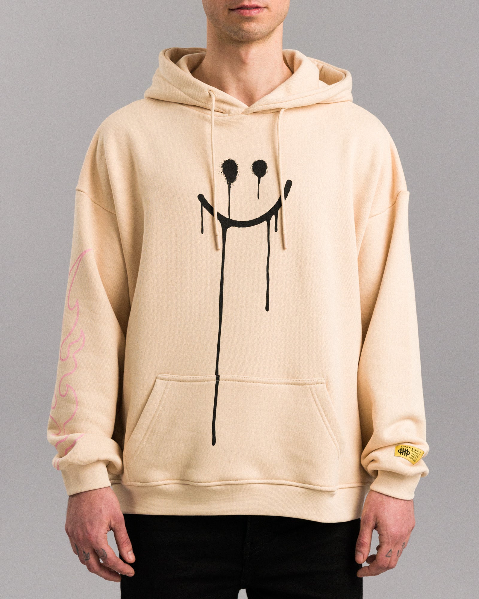 Beigefarbener Hoodie mit schwarzem Spray-Print eines tropfenden Smileys auf der Vorderseite, getragen von einer Person. Der Hoodie besitzt eine Kängurutasche und eine Kapuze mit Kordeln.