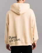 Rückansicht des beigefarbenen Hoodies mit schwarzem Schriftzug „RUNNING WITH SCISSORS“ im Graffiti-Stil am unteren rechten Saum. Die Kapuze liegt flach auf dem Rücken.