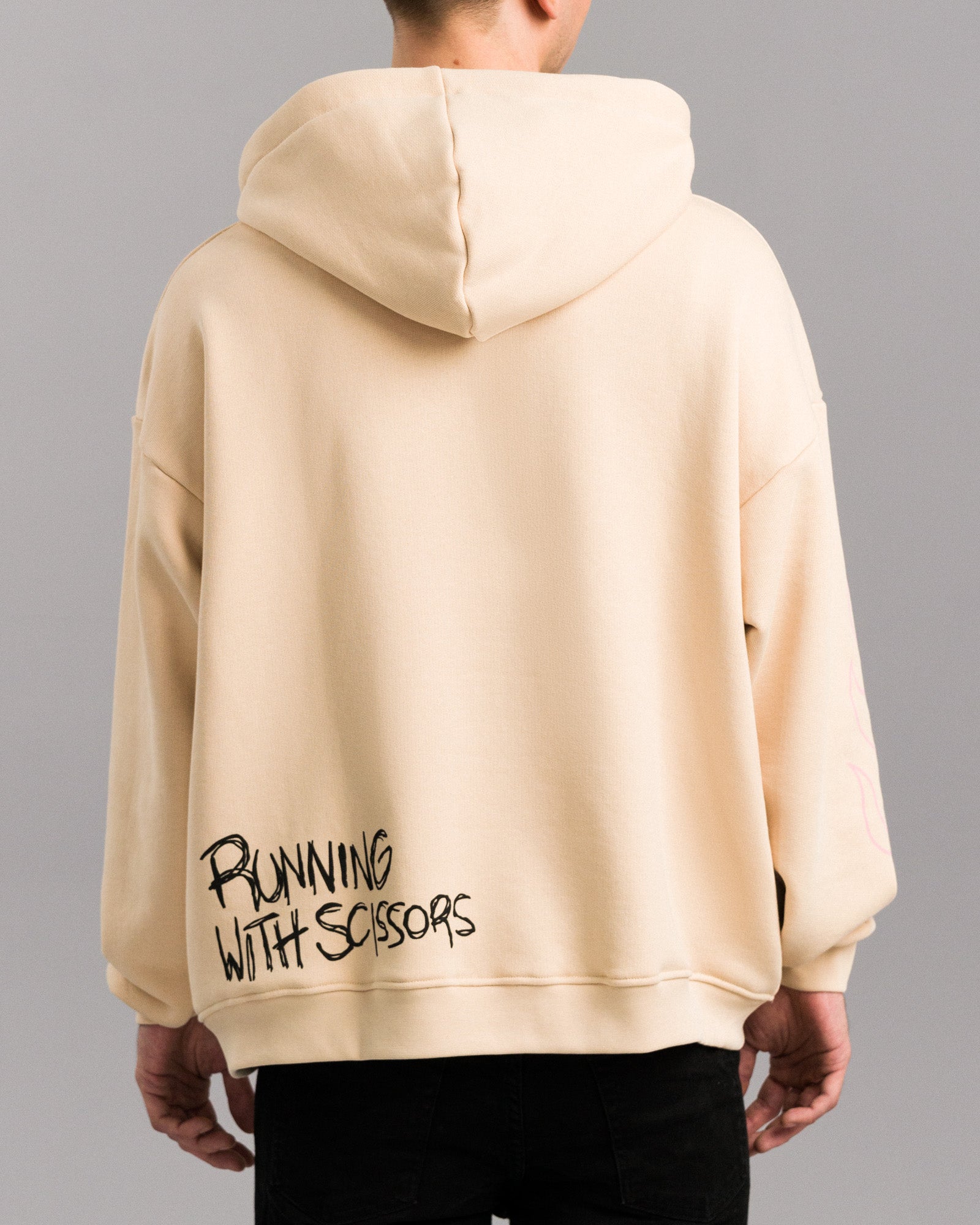 Rückansicht des beigefarbenen Hoodies mit schwarzem Schriftzug „RUNNING WITH SCISSORS“ im Graffiti-Stil am unteren rechten Saum. Die Kapuze liegt flach auf dem Rücken.