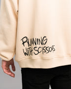 Detailansicht des Rückendrucks am Hoodie: Schwarzer Graffiti-Schriftzug „RUNNING WITH SCISSORS“ auf beigefarbenem Stoff, unten rechts platziert.